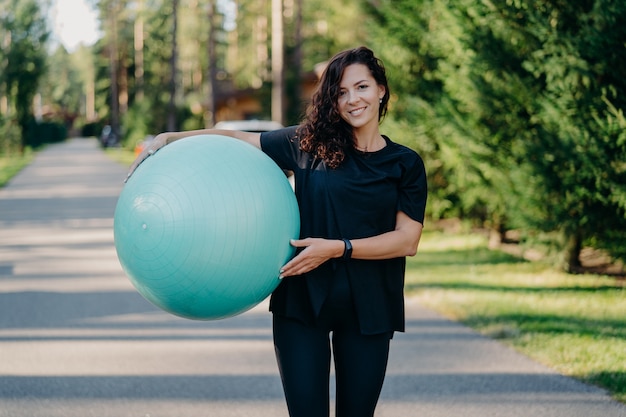 Colpo all'aperto di pose di donna bruna con fitball grande vestito con abiti sportivi ha formazione all'aria aperta