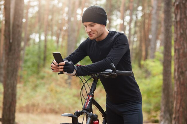 Colpo all'aperto orizzontale dello sportivo che posa intorno alla natura con la sua bicicletta, tenendo il suo telefono cellulare, toccando il suo schermo, utilizzando i siti di social network, avendo una breve pausa.