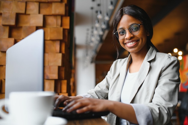 Colpo alla testa ritratto di felice sorridente donna afroamericana seduta al tavolo in caffè guardando la fotocamera eccitata femmina in posa lavorando al computer facendo i compiti preparando il rapporto in caffè
