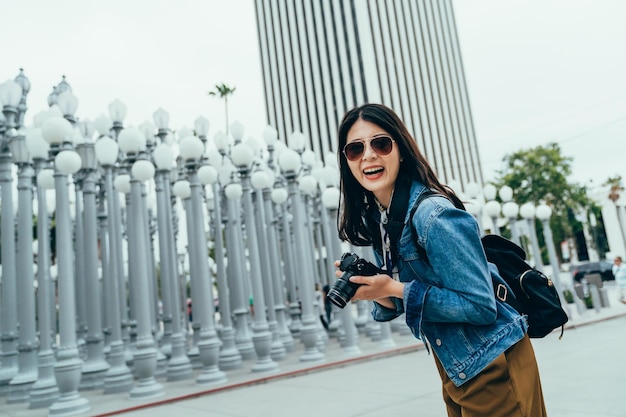 colpo d'angolo olandese La donna coreana che tiene la macchina fotografica digitale sta ridendo di cuore davanti a un'installazione artistica a Los Angeles. la turista asiatica che indossa occhiali da sole sta guardando l'obiettivo della fotocamera con un grande sorriso.