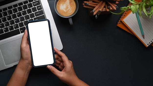 Colpo dall'alto delle mani della donna che tengono il telefono cellulare con lo schermo vuoto sul moderno posto di lavoro scuro.