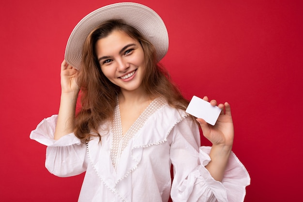 Colpo del primo piano della donna bionda scura adulta sorridente positiva attraente che indossa camicetta bianca e cappello bianco isolato sopra priorità bassa rossa che tiene la carta di credito che guarda l'obbiettivo.