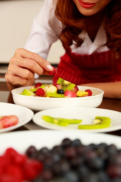 Colpo del primo piano della mano dello chef che mette il lampone in una deliziosa macedonia di frutta mista dolce in una ciotola bianca sul tavolo della cucina. Concetto di cibo biologico e naturale.