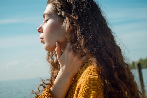 Colpo del primo piano di bella giovane donna con capelli ricci che porta in maglione giallo su una spiaggia, fuoco selettivo