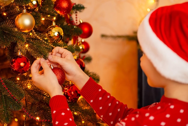 Colpo del primo piano di un ragazzino in cappello rosso della Santa che decora l'albero di Natale con le bagattelle che celebrano il Natale