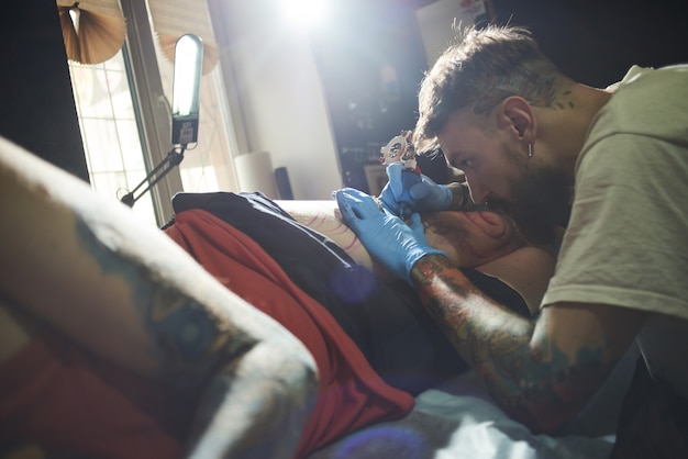 Colpo del primo piano di un tatuatore barbuto professionista che tatua un cliente ad un uomo in un salone