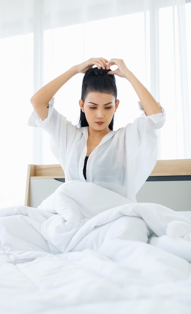 Colpo del ritratto della donna sexy asiatica millenaria in canotta comfort casual e camicia seduta sotto una coperta calda spessa bianca in posa braccio allungante dopo essersi svegliata in camera da letto la mattina a casa