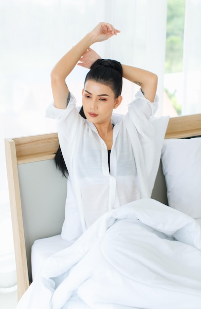 Colpo del ritratto della donna sexy asiatica millenaria in canotta comfort casual e camicia seduta sotto una coperta calda spessa bianca in posa braccio allungante dopo essersi svegliata in camera da letto la mattina a casa