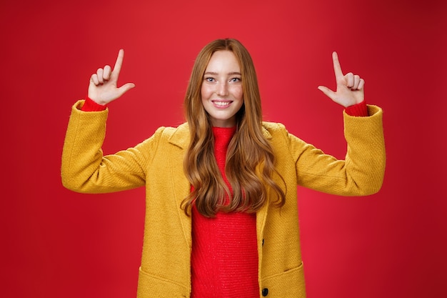 Colpo dell'interno della donna attraente della testarossa in cappotto giallo di caduta che alza le mani che promuovono la pubblicità come indicando su e che sorride ampiamente con l'espressione piacevole soddisfatta sopra fondo rosso. Copia spazio