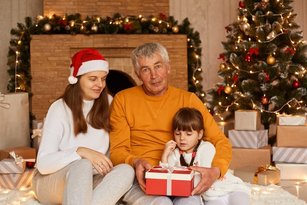 Colpo dell'interno della famiglia felice che dà il regalo di Natale alla piccola ragazza carina dai capelli scuri in una confezione regalo avvolta in rosso, in posa in un soggiorno decorato vicino all'albero di Natale e al camino.