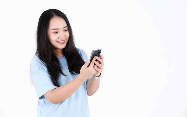 Colpo dello studio del modello femminile asiatico paffuto grassoccio lunghi capelli neri in maglietta casual blu e sorridente che tiene smartphone su priorità bassa bianca.