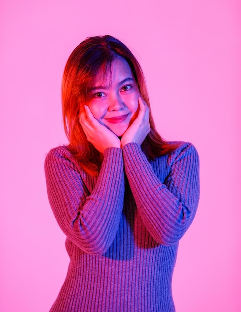 Colpo dello studio del ritratto di modello femminile asiatico giovane adolescente bella timido capelli tinti in maglia a collo alto manica lunga camicia in piedi in posa tenendosi per mano sulle guance guarda la macchina fotografica su sfondo rosa chiaro.