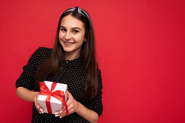 Colpo di affascinante ragazza bruna sorridente felice isolata su un muro di fondo rosso che indossa una camicetta nera che tiene in mano una scatola regalo bianca con un nastro rosso e che guarda l'obbiettivo. Copia spazio