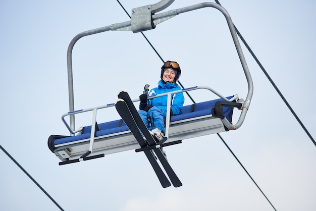 Colpo di angolo basso di uno sciatore femminile sorridente in tuta da sci blu in sella alla cima della montagna su un impianto di risalita