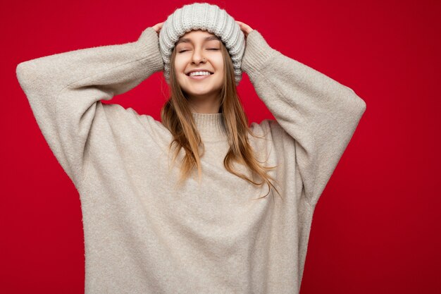 Colpo di attraente felice sorridente giovane donna bionda scura in piedi isolata sopra il muro di fondo rosso indossando maglione caldo beige e cappello beige invernale e godendo con gli occhi vicini.