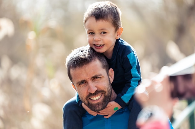 Colpo di bel giovane padre con il suo piccolo figlio sulle spalle guardando la telecamera mentre scattano una foto all'aperto.