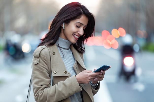 Colpo di bella giovane donna che ascolta la musica con gli auricolari wireless e lo smartphone in strada.