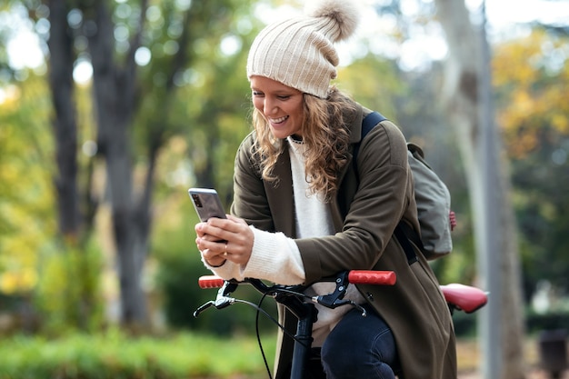 Colpo di bella giovane donna che invia messaggi con il suo smartphone mentre pedala attraverso il parco in autunno.