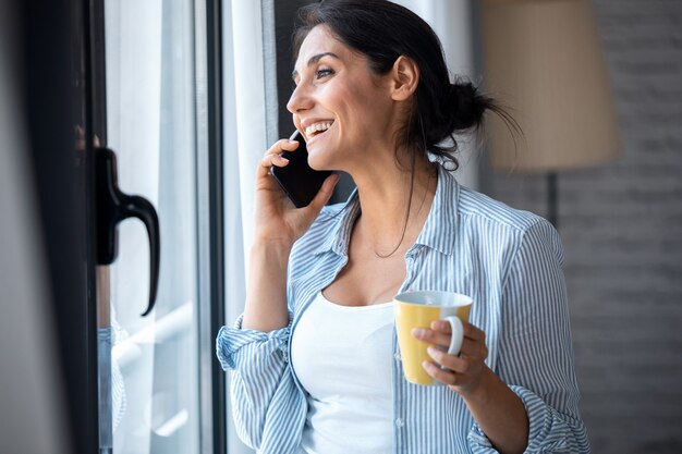 Colpo di bella giovane donna che parla al cellulare mentre beve caffè vicino alla finestra nel soggiorno di casa.