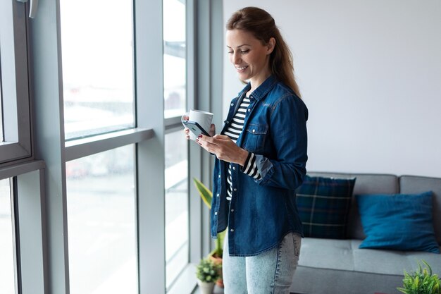 Colpo di bella giovane donna che usa il suo telefono cellulare mentre beve caffè vicino alla finestra nel soggiorno di casa.