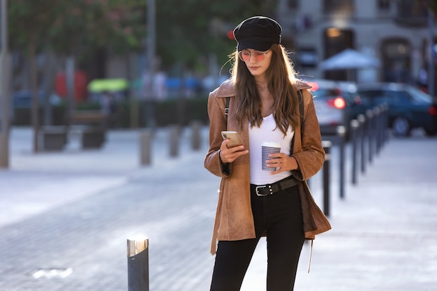 Colpo di bella giovane donna che usa il suo telefono cellulare mentre tiene in mano un caffè e sta in piedi per strada.