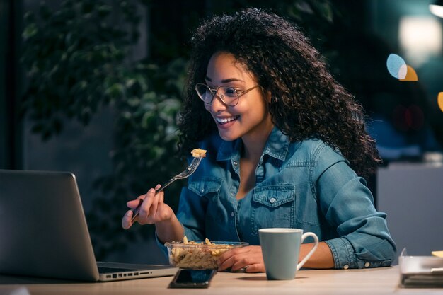 Colpo di bella giovane donna d'affari afro che lavora con il computer mentre mangia la pasta seduta in ufficio.