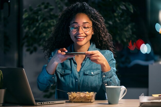 Colpo di bella giovane donna d'affari afro che scatta una foto con il suo smartphone mentre mangia pasta seduta in ufficio.