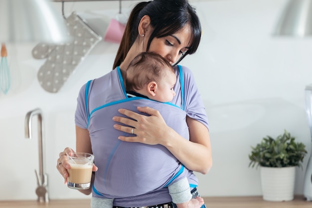 Colpo di bella giovane madre con bambino piccolo in fionda che beve caffè in cucina a casa.