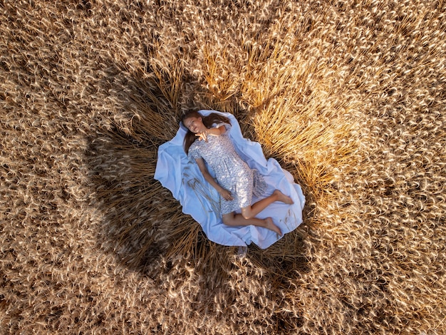 Colpo di drone dall'alto di una giovane ragazza sdraiata nel campo di grano maturo rivolto verso l'alto Bellissimo ritratto di una giovane ragazza nel campo di grano maturo