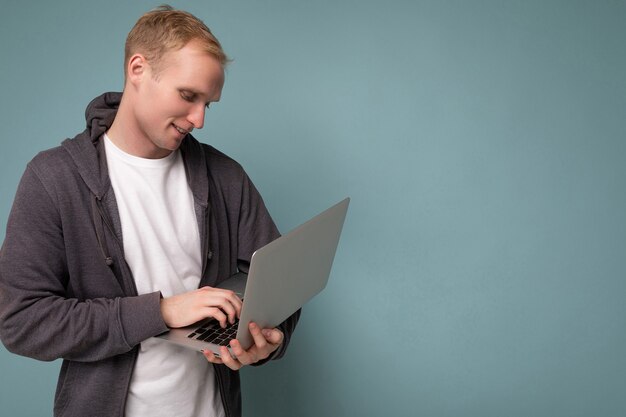 Colpo di foto del profilo laterale di bell'uomo biondo che tiene il computer portatile digitando sulla tastiera indossando t-shirt bianca e maglione grigio guardando netbook isolato su sfondo blu