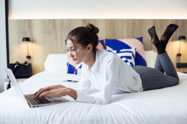 Colpo di giovane donna d'affari intelligente che lavora con il suo computer portatile sdraiato sul letto nella camera d'albergo.