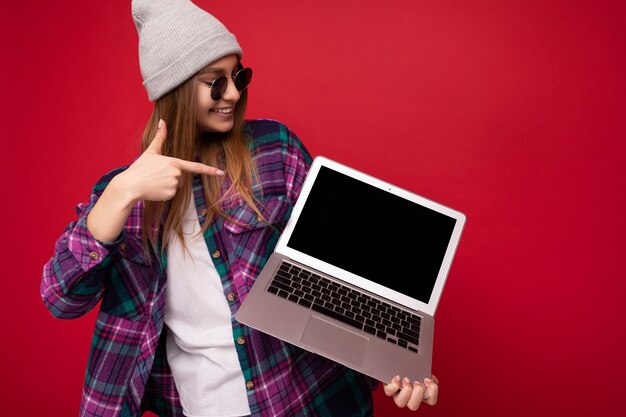 Colpo di giovane donna sorridente che tiene il computer portatile che indossa occhiali da sole cappello e maglietta colorata guardando il monitor del netbook e indicando il display del netbook isolato su sfondo rosso muro. Modello