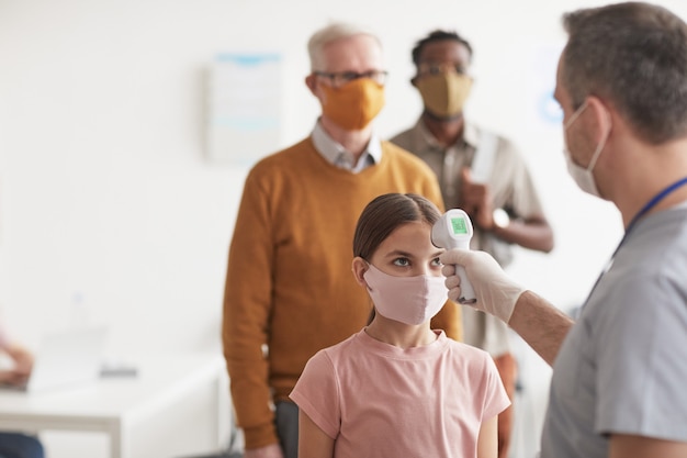 Colpo di medico irriconoscibile che controlla la temperatura della bambina che indossa la maschera e aspetta in fila in clinica o in ospedale, copia spazio