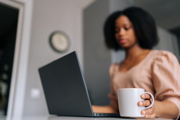 Colpo di messa a fuoco selettiva del primo piano di freelance femminile nera che digita sul laptop guardando lo schermo del dispositivo e tiene in mano una tazza con caffè o tè al mattino seduto al tavolo