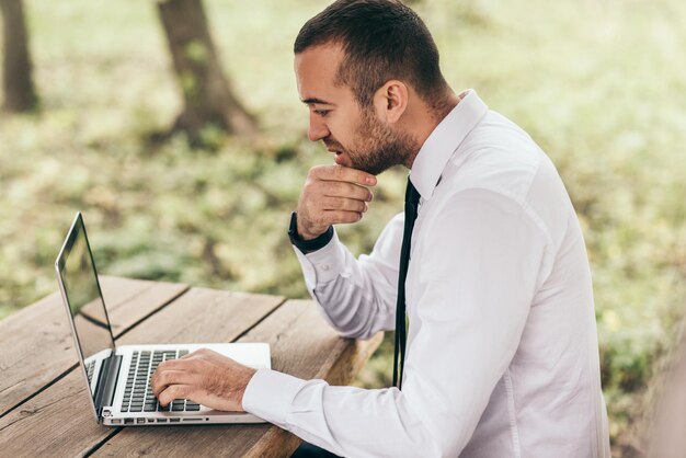 Colpo di profilo di un giovane uomo d'affari caucasico serio e concentrato che si sente frustrato seduto all'aperto con un pc portatile generico che tocca la barba cercando di concentrarsi sul lavoro che sembra esausto