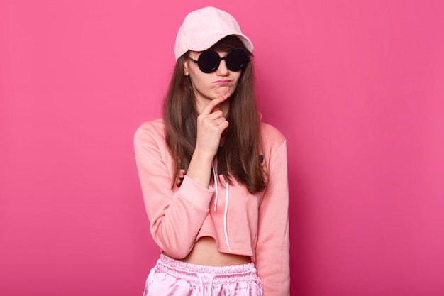 Colpo di Srusio della ragazza adolescente alla moda che indossa una felpa con cappuccio rosa accorciata alla moda, berretto e sotto-occhiali neri, le tocca il mento con un'espressione facciale ghettful. femmina. Concetto di persone.