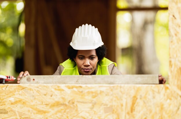 Colpo di un architetto femminile afroamericano che usa il livello sulla finestra mentre controlla il cantiere di una nuova casa di legno.
