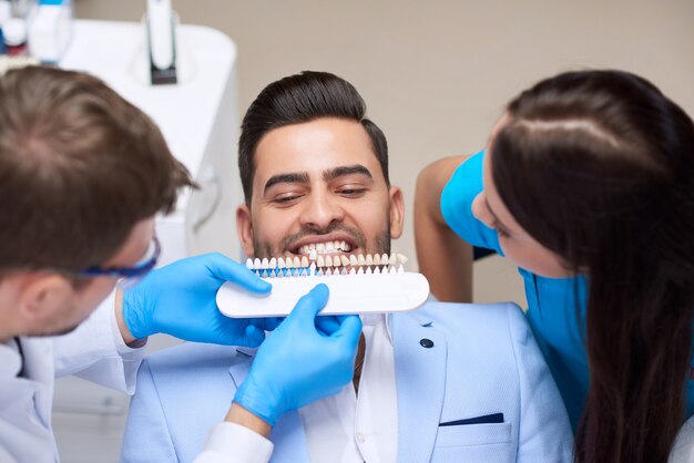 Colpo di vista dall'alto di un dentista professionista che wirking con l'assistenza dell'infermiere che sceglie il colore di corrispondenza perfetto degli impianti per le sue protesi dentarie sbiancanti per la medicina dei denti del paziente maschio.