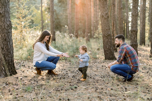 Colpo esterno orizzontale di bella giovane famiglia caucasica su una passeggiata nella foresta di autunno. Piccolo neonato bambino che cammina da suo padre a madre e ridendo