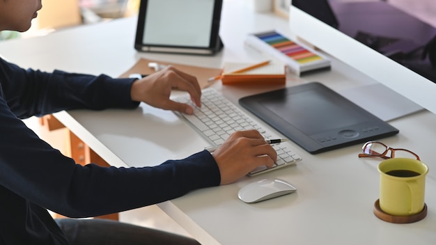 Colpo laterale di giovane uomo creativo mentre si digita sulla tastiera. Compresi tazza da caffè, mouse, tastiera, guida colori, auricolare tablet, taccuino e matita sulla scrivania.