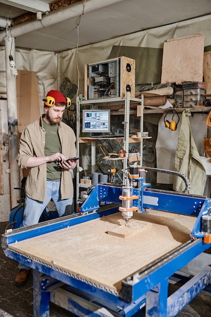 Colpo lungo verticale di un giovane uomo caucasico che usa il tablet durante il funzionamento di una fresatrice CNC in un'officina di lavorazione del legno