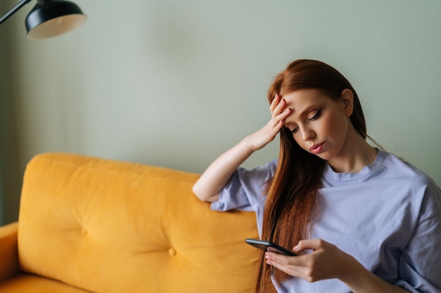 Colpo medio di giovane donna preoccupata in abiti casual seduta sul divano utilizzando il telefono a casa guardando lo schermo del gadget e toccando la testa pensando al problema