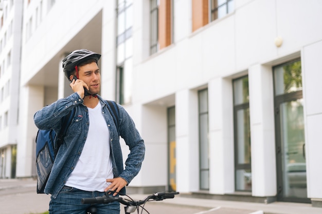 Colpo medio di un bel corriere maschio con zaino termico che parla al telefono cellulare con il cliente, cercando l'indirizzo per consegnare l'ordine online. Fattorino in casco protettivo utilizzando smartphone all'aperto.