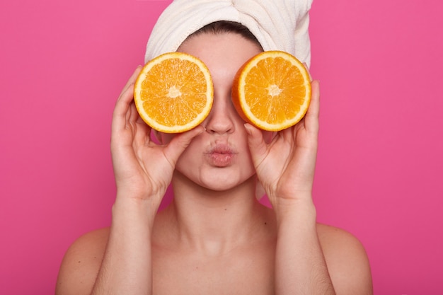 Colpo orizzontale della giovane donna allegra divertente che tiene due metà dell'arancia contro i suoi occhi, con l'asciugamano bianco sulla sua testa, posando con le spalle scoperte sopra la parete ottimistica. Concetto di bellezza.