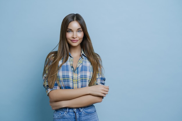 Colpo orizzontale di attraente giovane femmina con lunghi capelli lisci, indossa camicia a scacchi, jeans, tiene le mani incrociate, ha la pelle pulita, il trucco minimo, esprime emozioni positive, gode della sessione fotografica