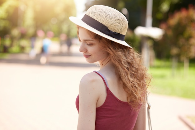 Colpo orizzontale di bella ragazza magra con i capelli ondulati in piedi nel parco tornando alla telecamera, indossando cappello di paglia, maglietta casual bordeaux, ha riposo con gli amici all'aria aperta, sembra romantico e gentile.