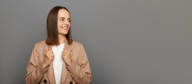 Colpo orizzontale di donna dai capelli scuri che indossa una giacca beige in piedi isolata su sfondo grigio guardando da parte lo spazio della copia per la pubblicità o il testo promozionale