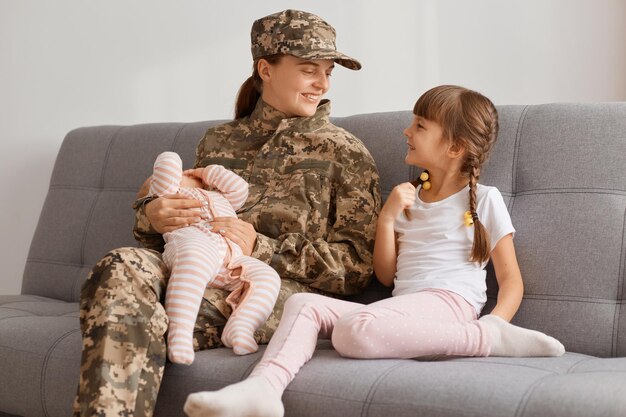 Colpo orizzontale di una donna soldato accattivante che indossa un'uniforme mimetica seduta sulla tosse con due figlie, che tiene in mano un neonato e parla con un bambino più grande.