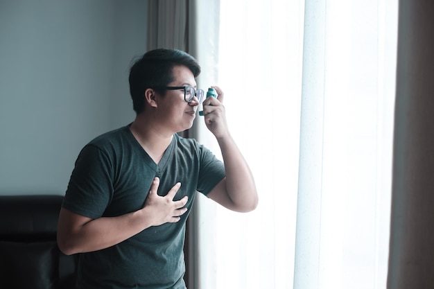 Colpo panoramico di uomo asmatico che usa l'inalatore con distanziatore Uomo grasso che usa l'inalatore per l'asma mentre è seduto a casa a causa dell'allergia Uomo asiatico che usa l'asma spray