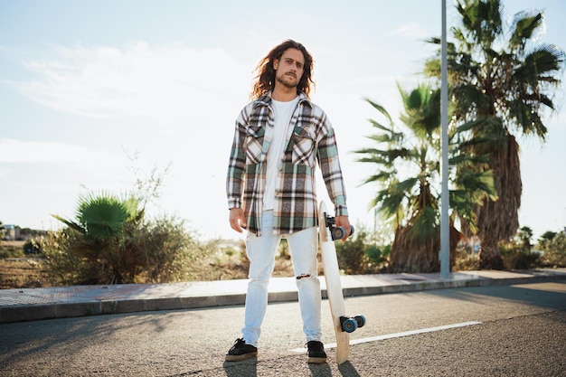 Colpo retroilluminato di un giovane serio che tiene il suo skateboard con palme sullo sfondo